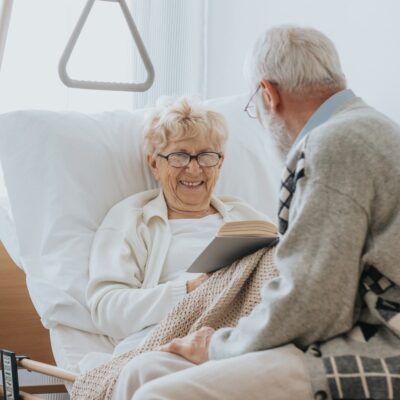 elderly woman in hospital bed holding a book and conversing with man sitting on the bed