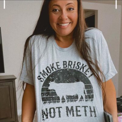 woman with long brunette hair smiling and showing off a "smoke brisket" shirt